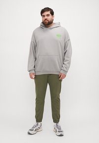 Hanorac gri cu buzunar frontal și detalii grafice verzi, asortat cu pantaloni tapered de un verde măsliniu și adidași gri cu accente albe.