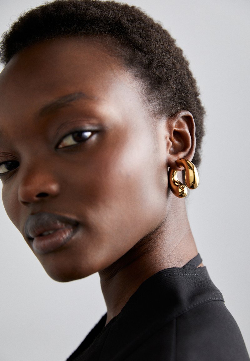 Gold-tone hoop earrings with a smooth, reflective surface. Worn on the ear, highlighting a curved and sculptural design.
