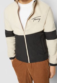 Fleecekangas- ja nailontakki, jossa beige yläosa ja musta alaosa, kokonapitallinen vetoketju, joustavat hihansuut ja kirjailtu logo.