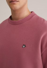 Prahasto roza pulover s ribanim okroglim izrezom in majhnim črnim vezenim logotipom ptice na levi prsni strani. Mehka tekstura.