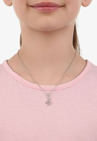 Collar de plata con un pequeño colgante de ciervo, con un brillo destacado, llevado sobre una camiseta de cuello redondo rosa claro. Cadena corta visible.
