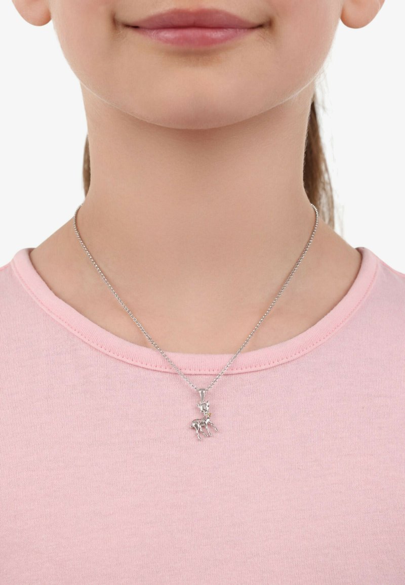 Silverhalsband med en liten hjortpresent, med en glänsande accent, bärs över en ljusrosa t-shirt med rund halsringning. Kort kedja synlig.