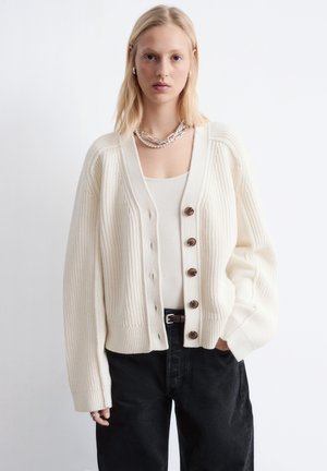 Blonde vrouw met een crèmekleurige gebreide cardigan met knopen over een witte top en zwarte broek, staand met één hand in de zak.