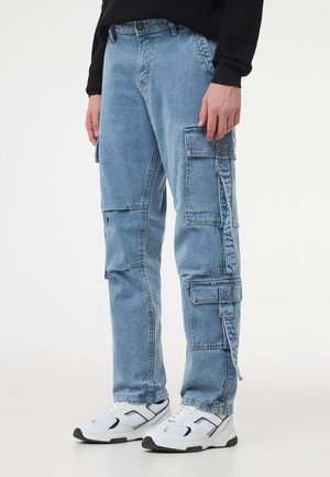 Pantaloni cargo in denim blu chiaro con stili di tasche multipli, design a gamba dritta, cuciture a contrasto e texture vissuta. Indossati con sneaker bianche.