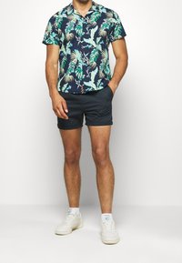 Krátký rukáv, námořnická košile s potiskem zelených tropických listů; v kombinaci s tmavými šortkami; bílé tenisky; ležérní střih.