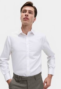 Weißes Hemd mit langen Ärmeln und klassischem Kragen aus glattem Stoff, kombiniert mit grauen, maßgeschneiderten Hosen.