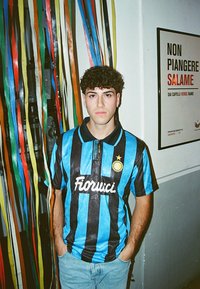 Blauwe en zwarte gestreepte voetbalshirt met "Fiorucci" logo, kraag en gouden ster. Draag met lichtblauwe jeans. Kleurrijke linten op de achtergrond.