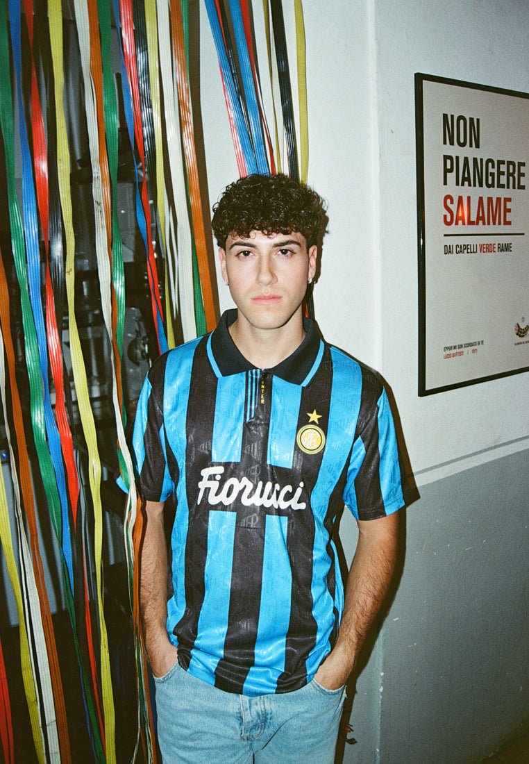 Blauwe en zwarte gestreepte voetbalshirt met "Fiorucci" logo, kraag en gouden ster. Draag met lichtblauwe jeans. Kleurrijke linten op de achtergrond.