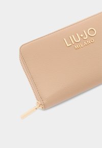 Beige leren portemonnee met een gestructureerde afwerking; voorzien van een gouden logo en ritsknop, met een schone, rechthoekige vorm.