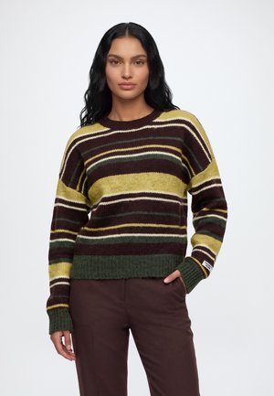 Une femme porte un pull rayé marron, jaune et vert, avec un pantalon bordeaux, des mocassins et un sac à bandoulière taupe, sur fond blanc.