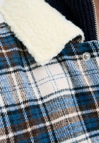 Veste en flanelle à carreaux avec des couleurs bleu, marron et crème ; col en sherpa blanc ; fermeture à pression argentée ; texture douce ; et intérieur côtelé.