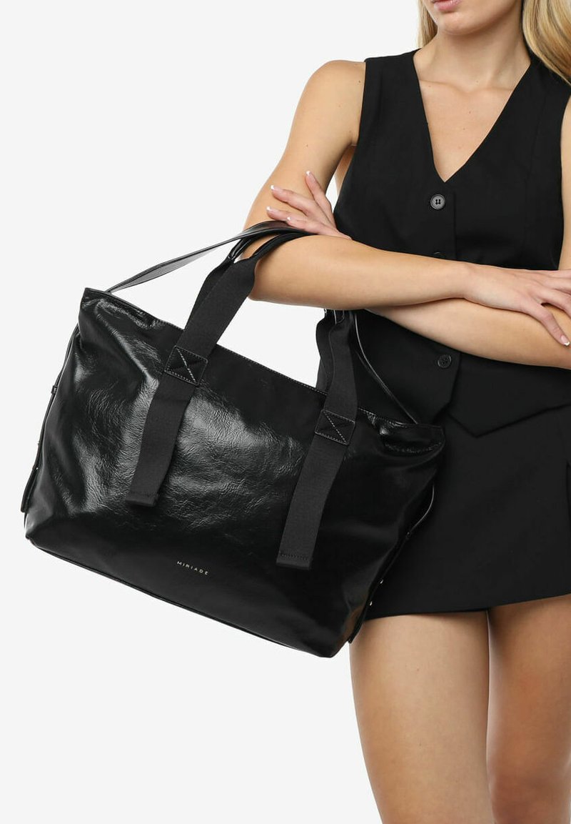 Zwarte leren tote bag met een gestructureerde afwerking, dubbele stoffen handvatten en een minimalistisch logo aan de onderkant. Een veelzijdig, ruim ontwerp.