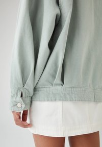 Veste oversize vert menthe en tissu texturé, avec des détails de poignets boutonnés et un ourlet simple, associée à une jupe blanche.