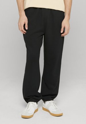 Mann, der schwarze elastische Bundhosen und weiße Turnschuhe trägt, steht vor einem schlichten Hintergrund.