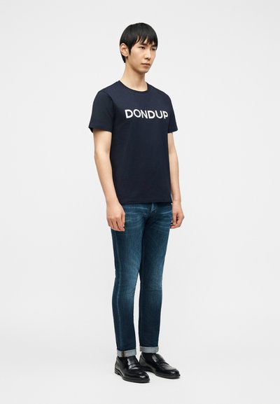 Άνδρας που στέκεται φορώντας μαύρο t-shirt με το λογότυπο "DONDUP", καλοκουμπωμένο μπλε τζιν, μαύρες κάλτσες και μαύρα δερμάτινα παπούτσια μπροστά σε απλό λευκό φόντο.