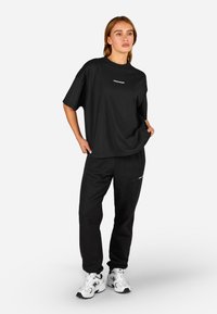 Μαύρη υπερμεγέθη μπλούζα με κοντά μανίκια, στρογγυλή λαιμόκοψη και λεπτομέρεια λογότυπου. Συνδυάζεται με μαύρες φόρμες και λευκά αθλητικά παπούτσια. Λεπτή υφή.