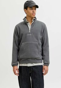 Pullover en polaire gris avec demi-fermeture éclair, poche avant et logo "JJ Studios" brodé. Porté par-dessus une chemise à carreaux et associé à un jean foncé.