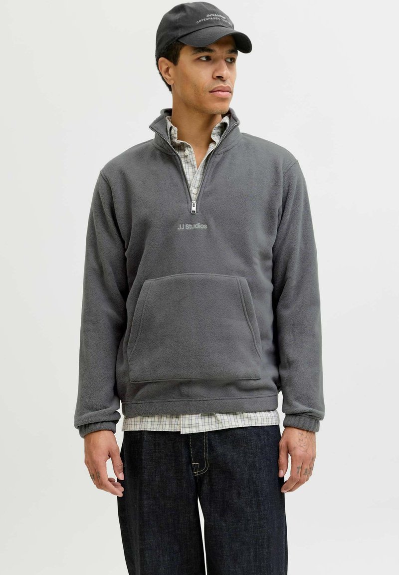 Pullover en polaire gris avec demi-fermeture éclair, poche avant et logo "JJ Studios" brodé. Porté par-dessus une chemise à carreaux et associé à un jean foncé.