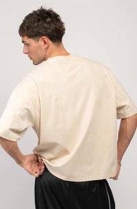 Man with short hair and earrings wearing a loose beige T-shirt and black shorts, posing with hands on hips, facing away.