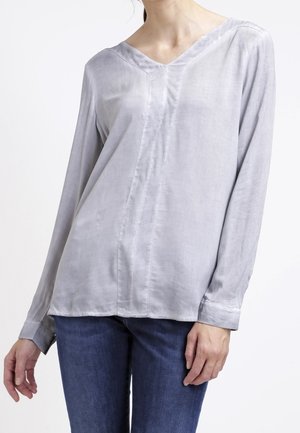 Femme portant un chemisier gris clair à manches longues avec col en V et un jean bleu, debout devant un fond blanc uni.