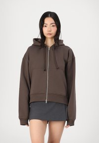 Sudadera con cremallera marrón de corte relajado, con grandes bolsillos frontales, capucha con cordón y puños de canalé. Lleva una falda gris oscuro debajo.