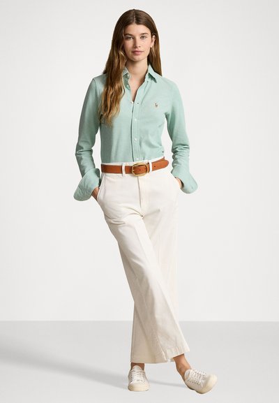 Camicia verde chiaro a maniche lunghe, pantaloni bianchi a gamba larga, cintura marrone e sneakers beige. Presenta un piccolo logo sulla camicia e le maniche arrotolate.