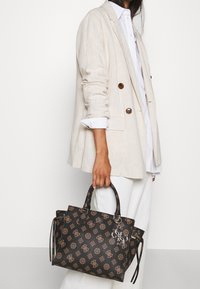 Sac fourre-tout marron à motif avec une forme structurée, des poignées en cuir et des éléments en métal argenté. Le design présente des motifs floraux complexes.