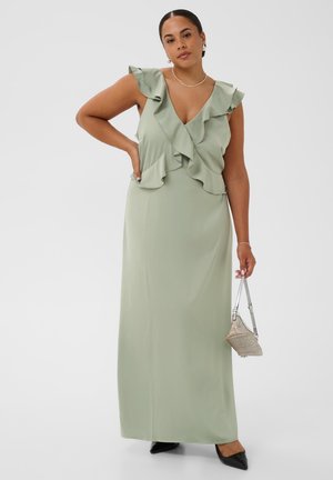 Femme portant une robe longue sans manches à volants vert clair, tenant une petite pochette argentée, debout avec une main sur la hanche.