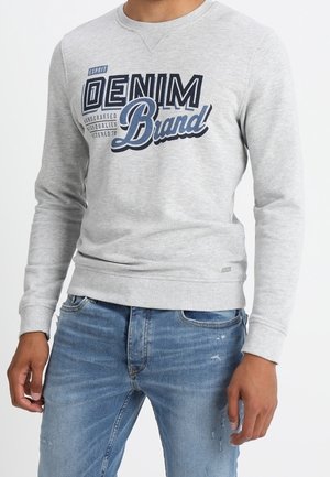 Hombre con sudadera gris claro con el logo "Denim Brand" en azul marino y azul, combinado con jeans desgastados de color azul claro, de pie contra fondo blanco.