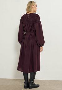 Vestito bordeaux con lunghe maniche a palloncino, corpetto arricciato e cintura in vita. Termina sotto il ginocchio, abbinato a stivali neri di pelle al ginocchio.