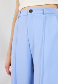 Světle modré kalhoty s širokými nohavicemi z hladké látky, s předním zapínáním na knoflík, bočními kapsami a ostrými záhyby.