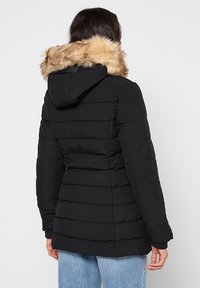 Doudoune noire avec capuche, dotée d'un col en fausse fourrure. Design texturé et matelassé avec des rayures. Portée par-dessus un jean bleu clair.