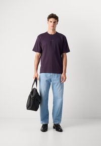 T-shirt en coton violet avec un col rond et des manches courtes, portée avec un jean en denim bleu clair et des chaussures en cuir noires, tenant un sac noir.