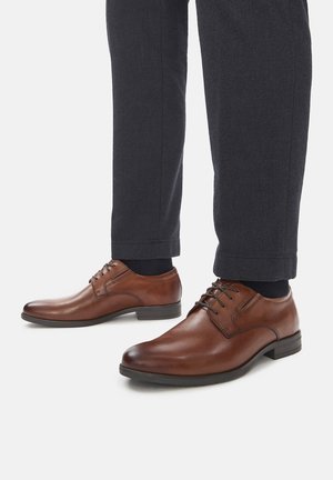 Brązowe skórzane eleganckie buty o gładkim wykończeniu, wyposażone w sznurowadła i niski obcas, zestawione z ciemnymi teksturowanymi spodniami i czarnymi skarpetkami.