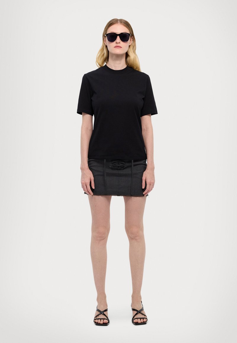 T-shirt nero a maniche corte abbinato a una mini gonna in denim nera con dettagli frontali e sandali neri con cinturini. Occhiali da sole scuri completano il look.