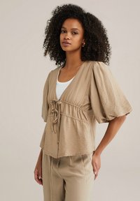 Kurzärmlige, beigefarbene Bluse mit Struktur, gerafftem Bund und einem Bindedetail. Kombiniert mit passenden Hosen, die einen einfachen runden Ausschnitt haben.