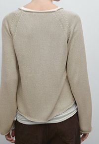 Beige gebreide trui met een textuurafwerking, raglanmouwen en een afgeronde zoom. Draagt over een witte gelaagde top.