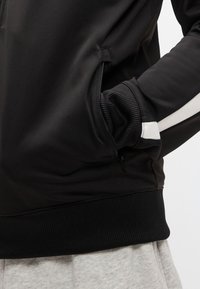 Veste de sport noire avec poignets et taille côtelés. Comprend une poche zippée et des bandes d'accent blanches sur les manches. Texture lisse.