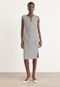 Robe midi à carreaux noir et blanc avec un décolleté en V, des manches courtes, une fente sur le côté et une taille ceinturée. Le modèle l'associe avec des talons blancs.