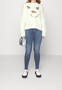 Helle Pastellfarbe Sweater mit einem verspielten Gesichtdesign, blaue Slim-Fit-Jeans und weiße Mary Jane-Schuhe mit schwarzen Sohlen, während eine silberne Handtasche gehalten wird.