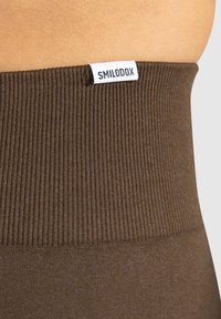 Brązowy prążkowany pas legginsów z białą metką z logo "SMILODOX". Materiał wydaje się gładki i elastyczny.