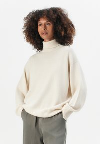 Cremfarbener Rollkragenpullover mit lockerer Passform, überschnittenen Schultern und gerippten Abschlüssen. Kombiniert mit hellgrauen Hosen aus weichem Material.
