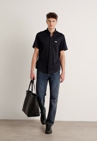 Camicetta nera a maniche corte con bottoni e logo ricamato, abbinata a jeans blu scuri e con una borsa a tracolla nera. Scarpe nere completano il look.
