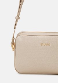 Borsa a tracolla beige in materiale tessurizzato con chiusura a zip. Presenta hardware color oro e logo del brand impresso sulla parte anteriore.