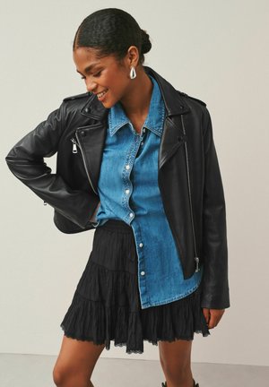 Žena sa usmieva, nosí strieborné kruhové náušnice, čiernu koženú bundu cez modrú džínsovú košeľu na gombíky a čiernu viacvrstvovú sukňu.