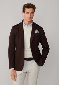 Mann trägt dunkelbraunen Blazer, cremefarbenen Pullover, hellblaues Hemd, hellfarbene Hose und ein gemustertes Einstecktuch, steht vor schlichtem Hintergrund.
