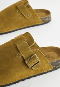 Brune ruskinds slip-on sko har en justerbar rem med en metalspænde, en polstret indersål og en tekstureret sort gummisål.
