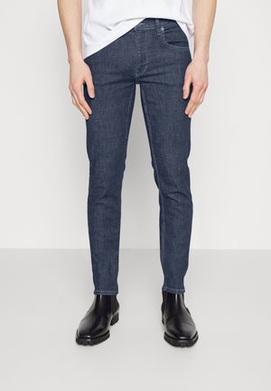Jeans de mezclilla azul oscuro de corte ajustado, con una cintura de media altura, cinco bolsillos y una ligera textura con costuras en contraste. Combinados con botas negras.