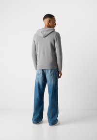 Mann som står med ryggen til, iført en grå hettegenser, blå slitte vide jeans og hvite joggesko, mot en enkel hvit bakgrunn.
