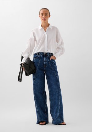 Weißes Hemd mit langen Ärmeln, kombiniert mit hoch taillierten, weiten blauen Jeans. Die Person hält eine schwarze Handtasche.